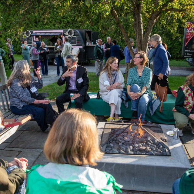 A group of people sitting around a fire pit in an outdoor setting, talking and engaging with one another. Trees, a food truck, and other attendees are visible in the background.