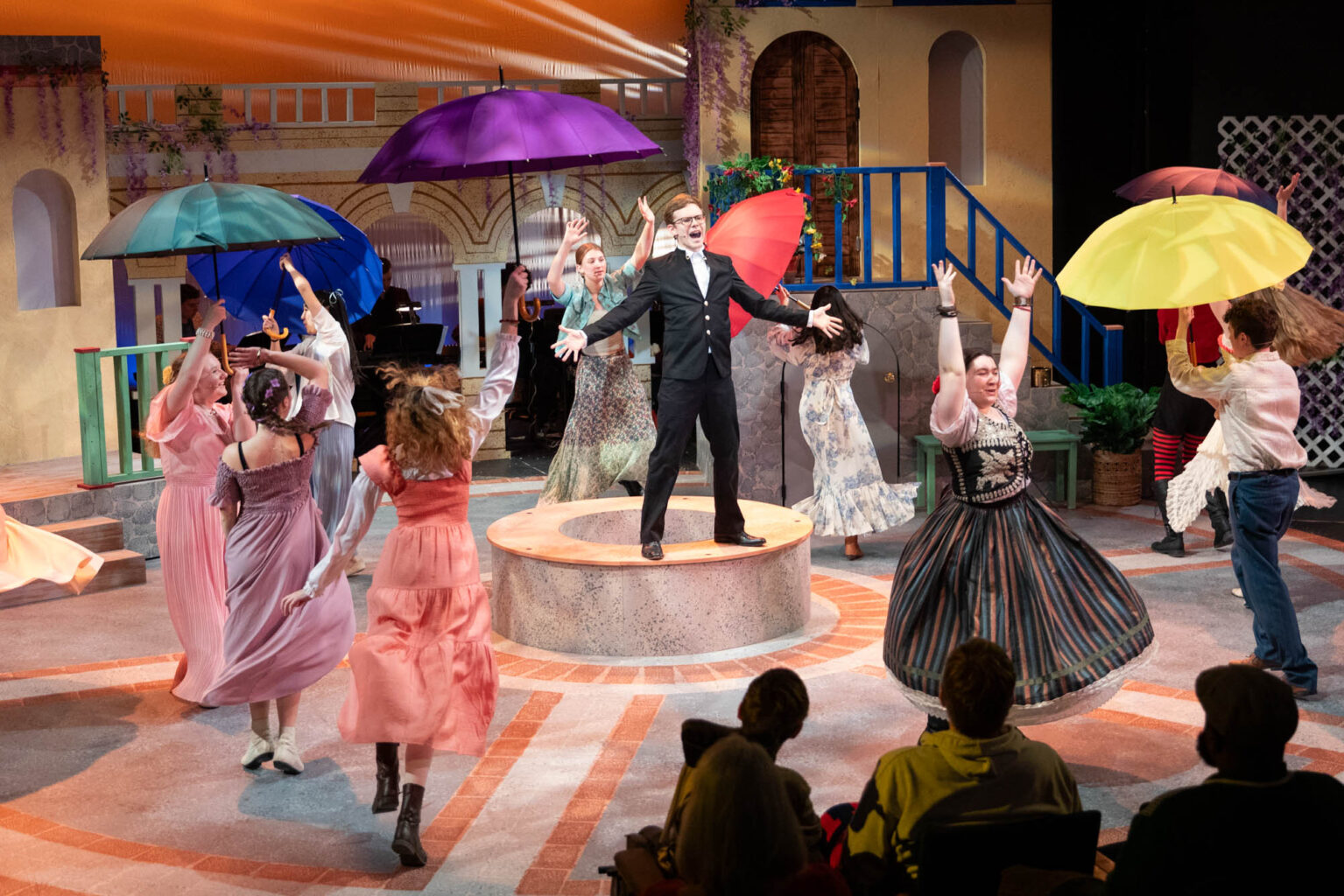 A theatrical performance with actors in colorful costumes dancing around a man in a suit standing on a circular platform. Some actors are holding vibrant umbrellas. An audience watches from the foreground.