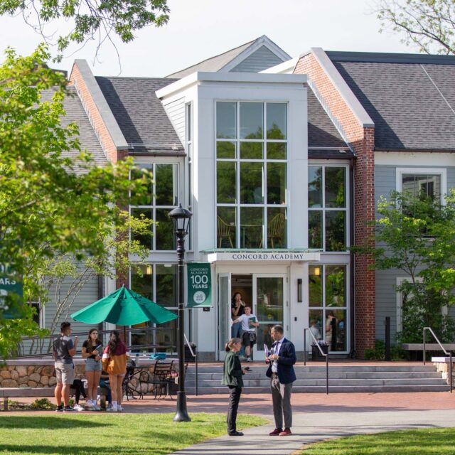 A group of people stand and sit near the entrance of a building labeled "Concord Academy" surrounded by greenery on a sunny day.