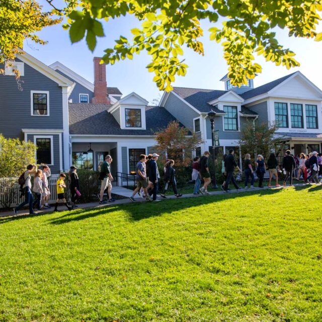 A group of people walking along a pathway beside a row of houses, with trees and green grass in the foreground on a sunny day.