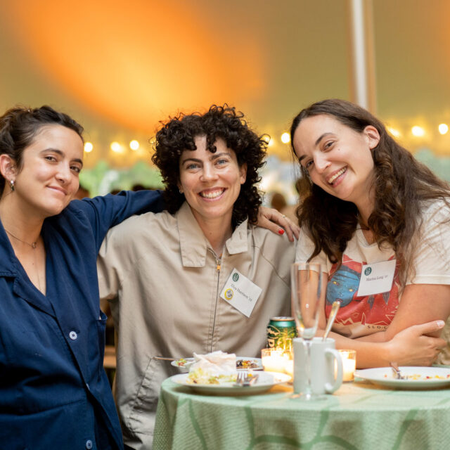 Three women smile at the camera while standing around a table at an outdoor event. The table holds plates, cups, and a can. String lights are visible in the background.