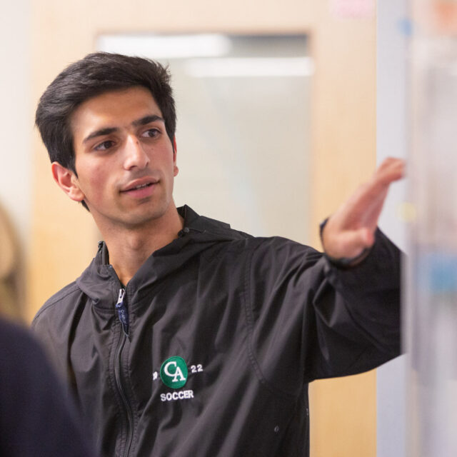 A young man in a jacket points at something off-camera while a woman in the foreground looks in a different direction. The setting appears to be indoors, possibly in a school or office.