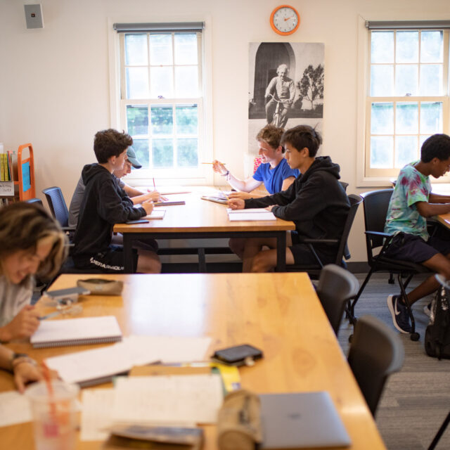 Students are seated at tables in a classroom, working on assignments. Two windows and a clock are on the back wall, along with a large black-and-white poster of a person.