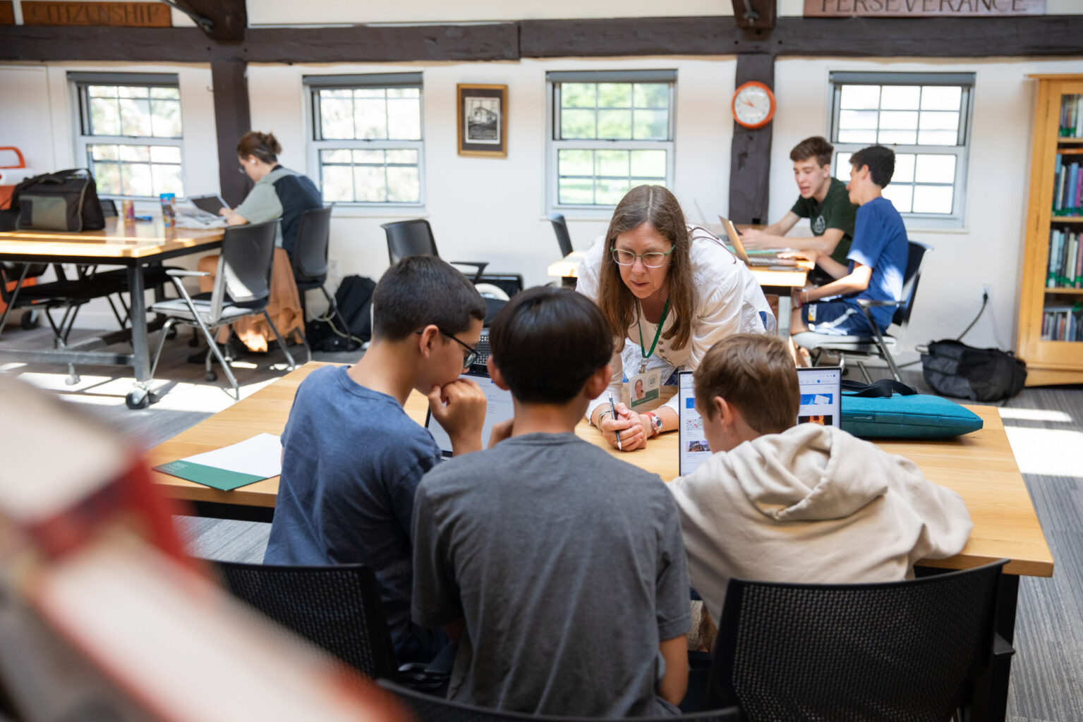A group of students and a teacher are engaged in a discussion around a laptop in a classroom with wooden beams and bookshelves. Other students are visible working at separate tables in the background.