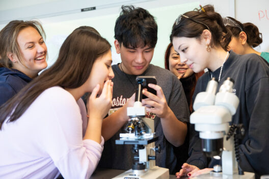 A group of students huddled around a smartphone in a science classroom, smiling and discussing something. Two microscopes are on the table in front of them.