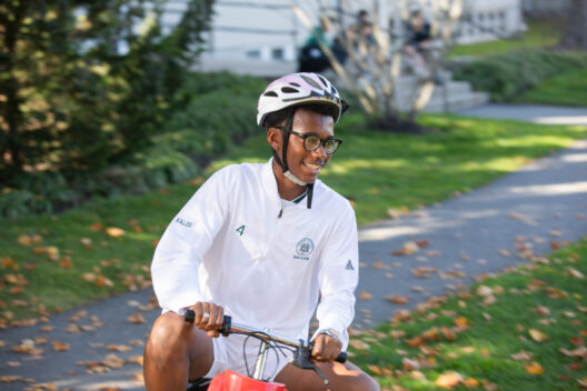 Person wearing a white helmet and white jacket, riding a bicycle on a pathway in a park-like setting with green grass and scattered fallen leaves.
