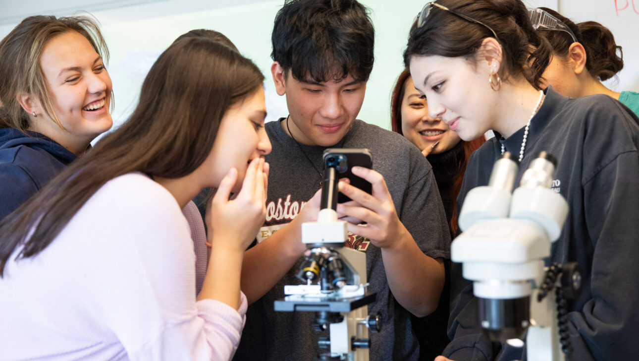 A group of students gathers around a microscope in a classroom, looking at a phone held by one student.