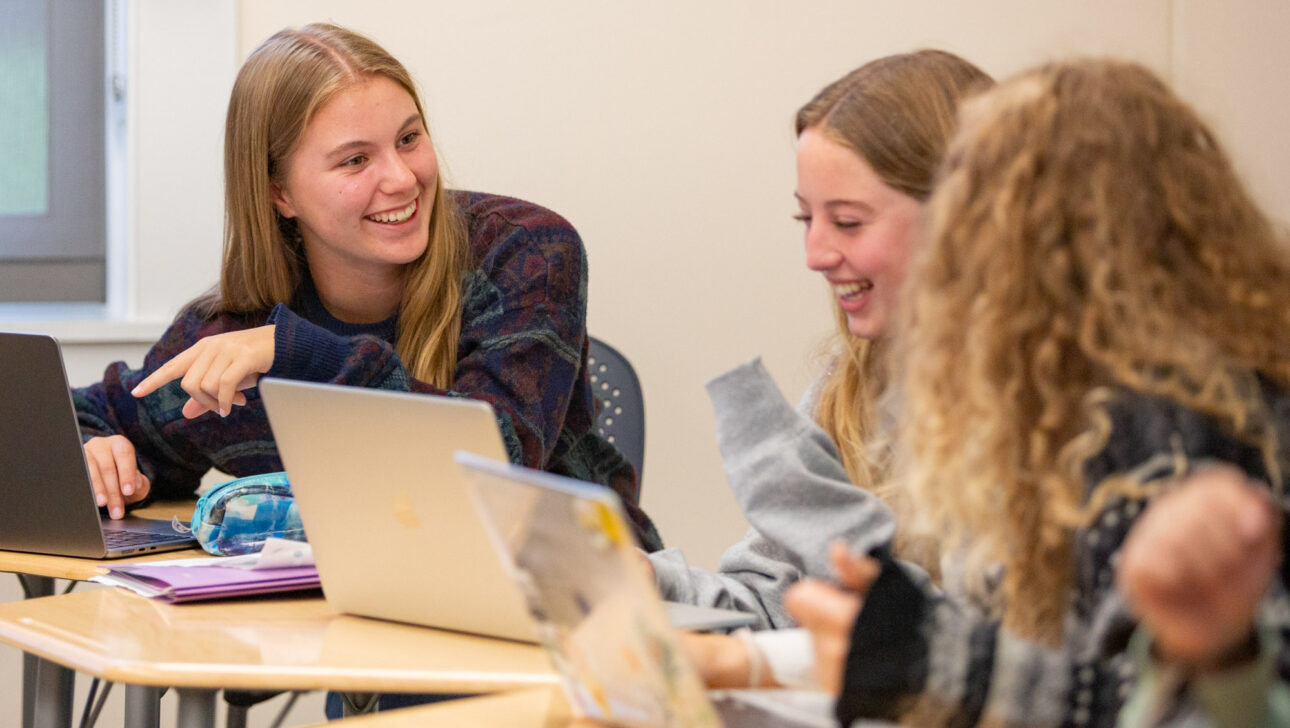 Three young women sit at desks using laptops, with one pointing and smiling at another. They appear to be engaged in a friendly conversation in a classroom setting.