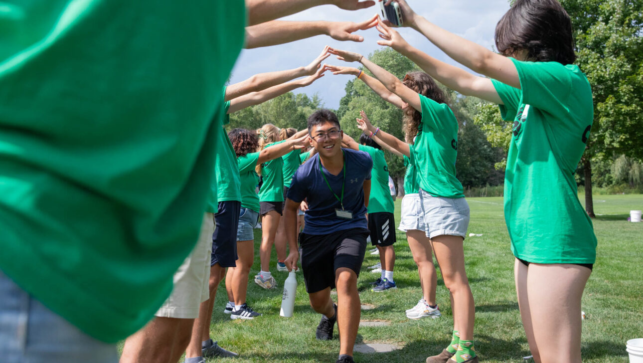 A person in a blue shirt runs through a tunnel formed by people in green shirts standing on a grassy field.