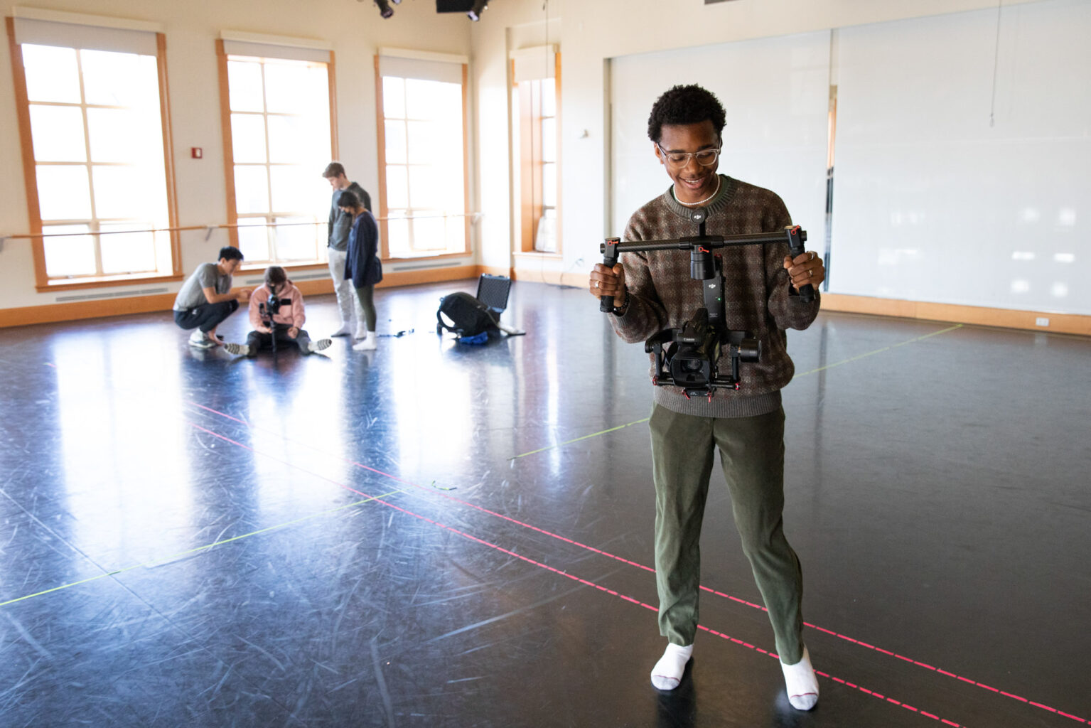 A person stands holding a camera stabilizer in a large room, while three others are together at the far end of the room near a window.