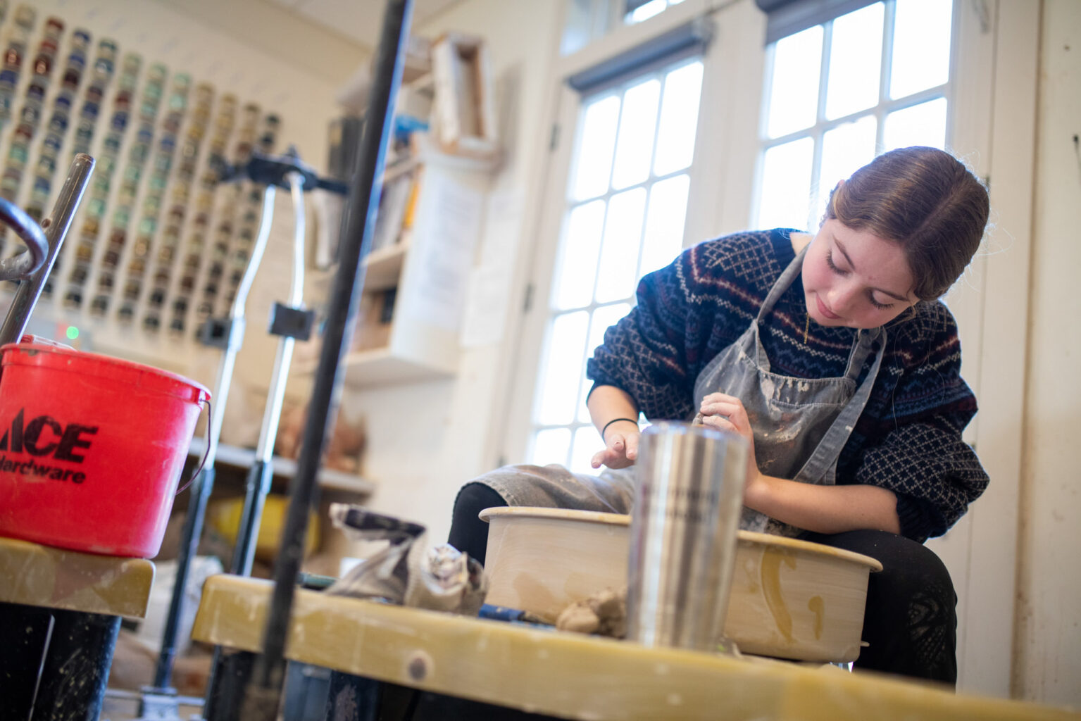 A person wearing a sweater and apron works on a pottery wheel in a well-lit studio with various tools and materials around.