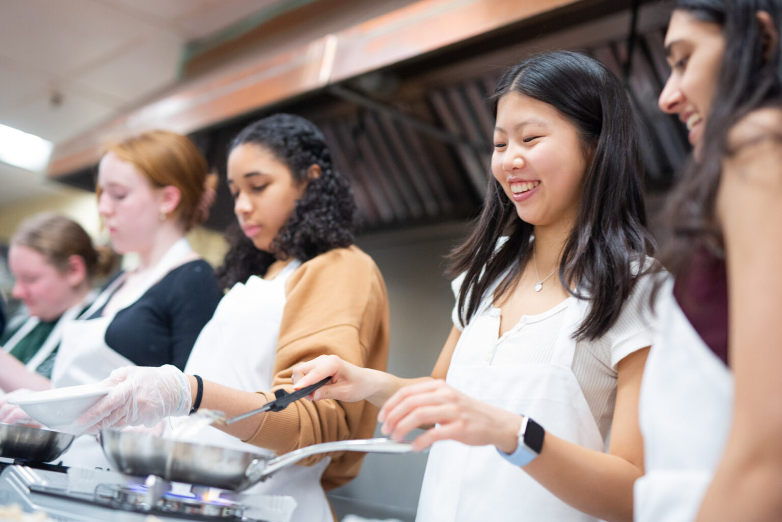 A group of five women wearing aprons and cooking in a kitchen. One woman in the center is smiling and holding a frying pan.