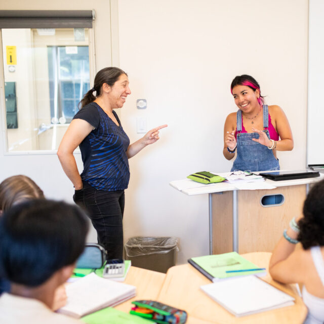 A teacher and student at the front of a classroom speaking to one another.