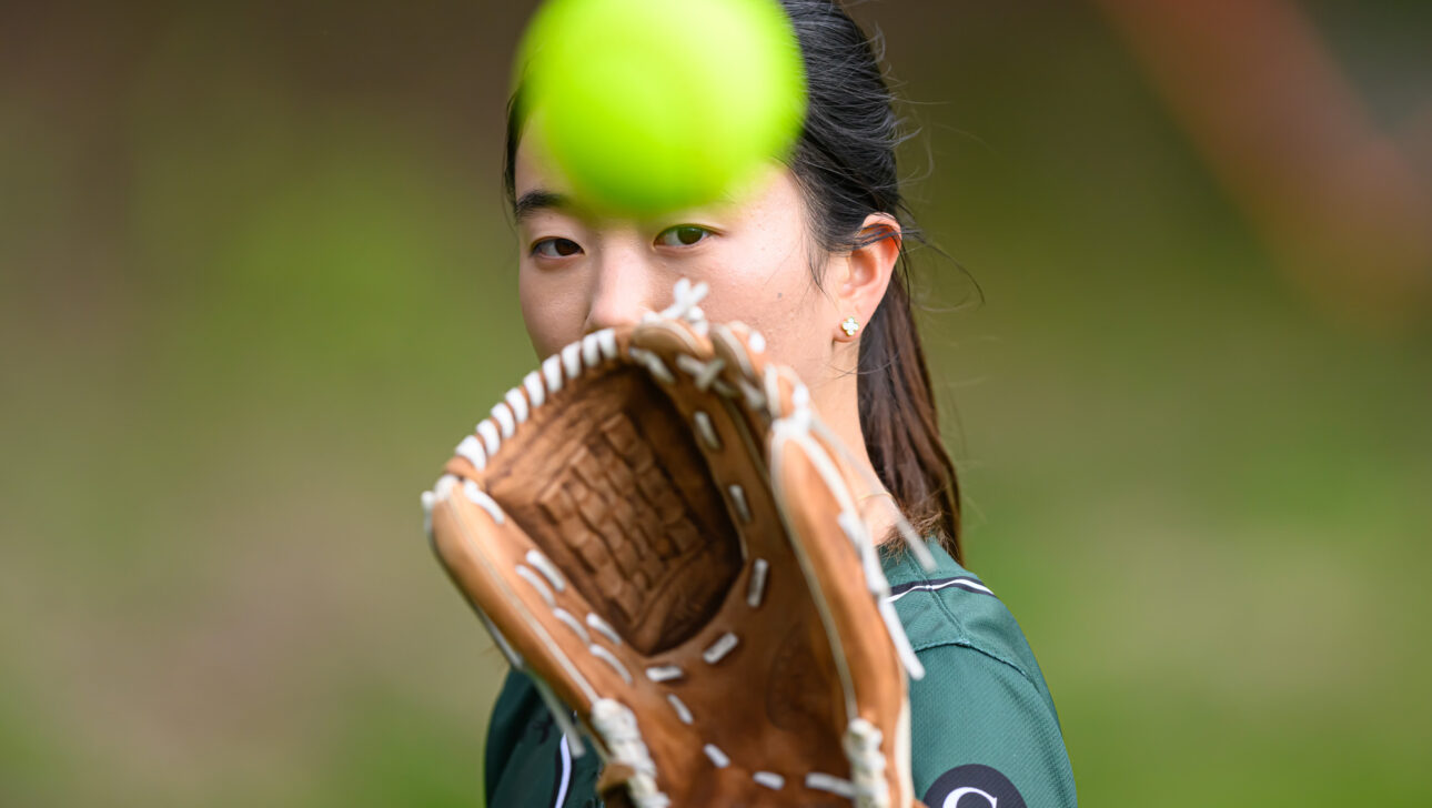 A person wearing a green shirt catches a yellow softball with a brown glove. The focus is on the ball and glove, with the person's face partially visible in the background.