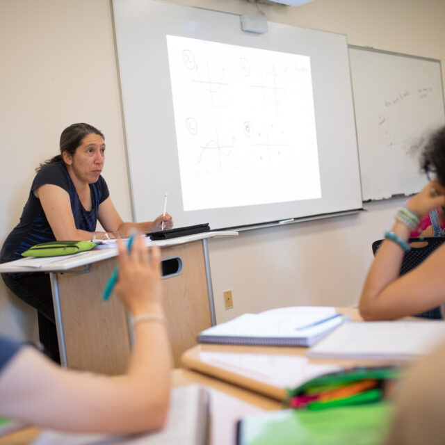 A teacher stands at a podium in a classroom, pointing at a projection of graphs on the wall. Students listen and take notes.