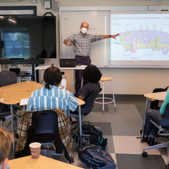 A professor, wearing a mask, stands at the front of a classroom pointing to a slide about cell membranes with students seated at round tables observing.