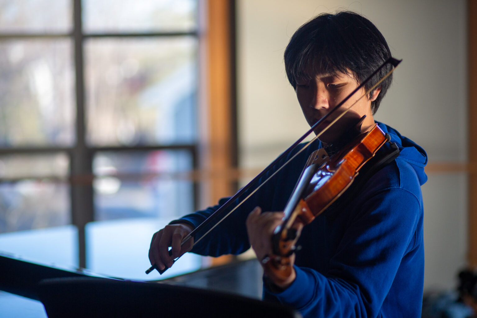 Person playing a violin by a window, wearing a blue jacket. The background is softly lit with natural light coming through the window.