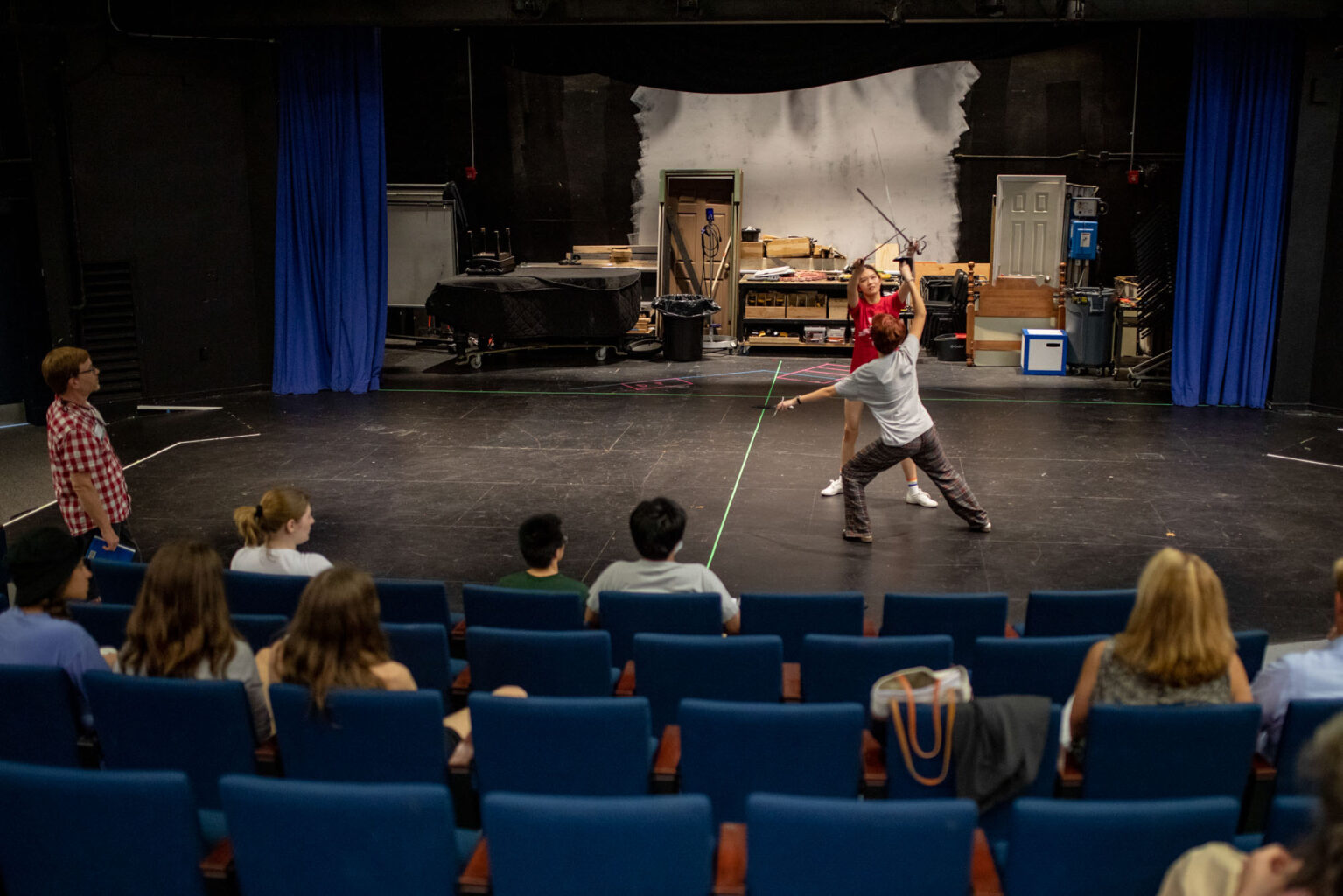 A group of people sits in theater seats watching two individuals on stage, one holding a sword, while participating in a rehearsal or practice session.