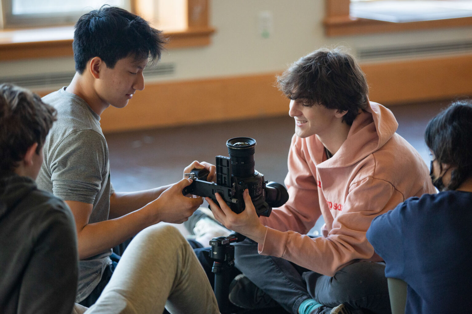 Four individuals are seated on the floor, examining a professional camera, with one person holding and pointing at it while another person listens attentively.