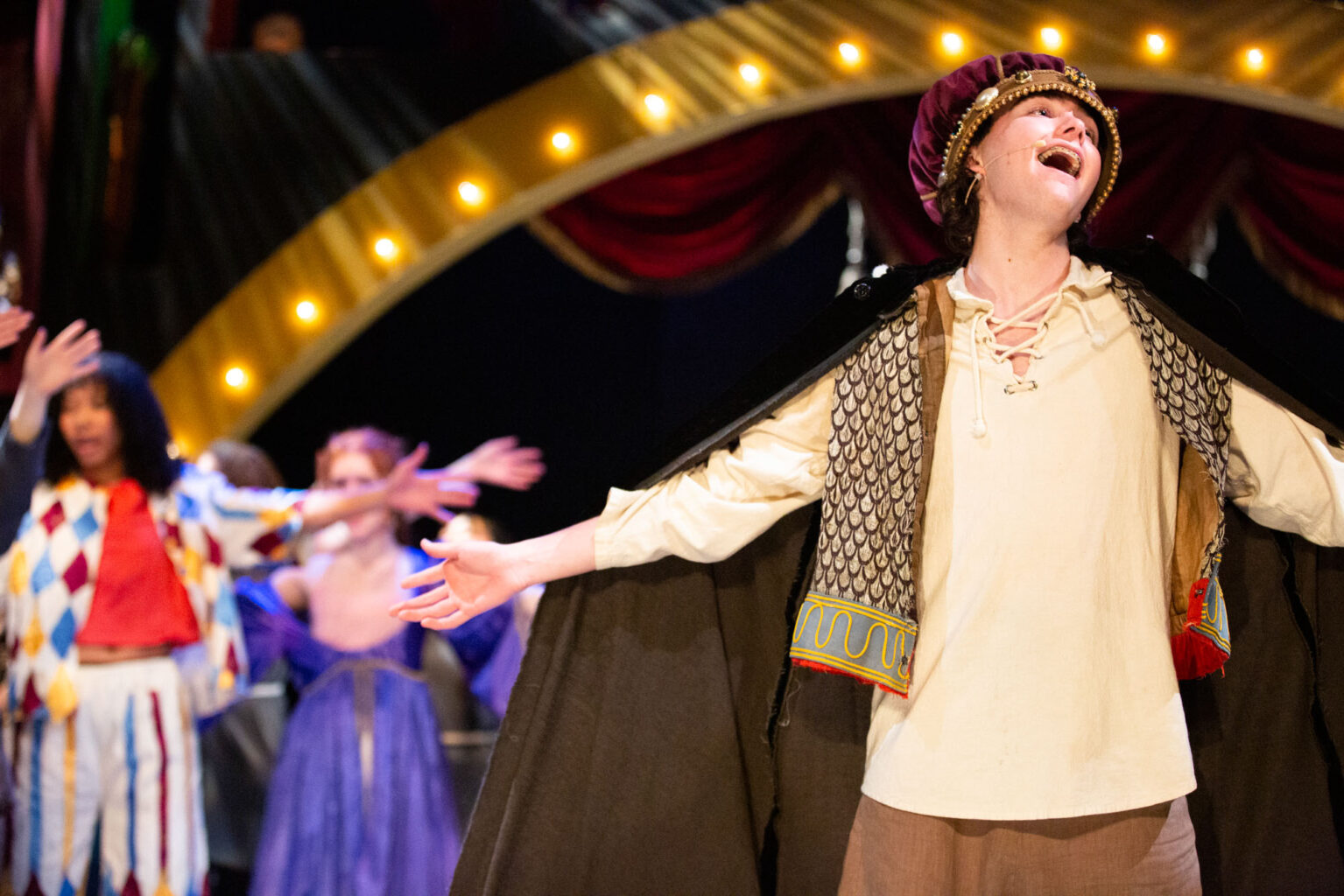 A performer dressed in medieval clothing sings passionately on stage, with other performers and a decorative backdrop featuring lights and red drapes in the background.