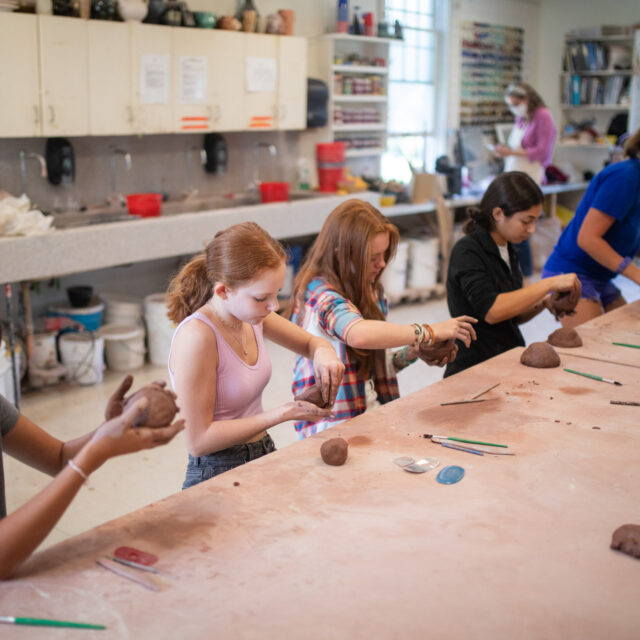 A group of people sitting at a long table, each working with clay in an art studio filled with supplies and equipment.