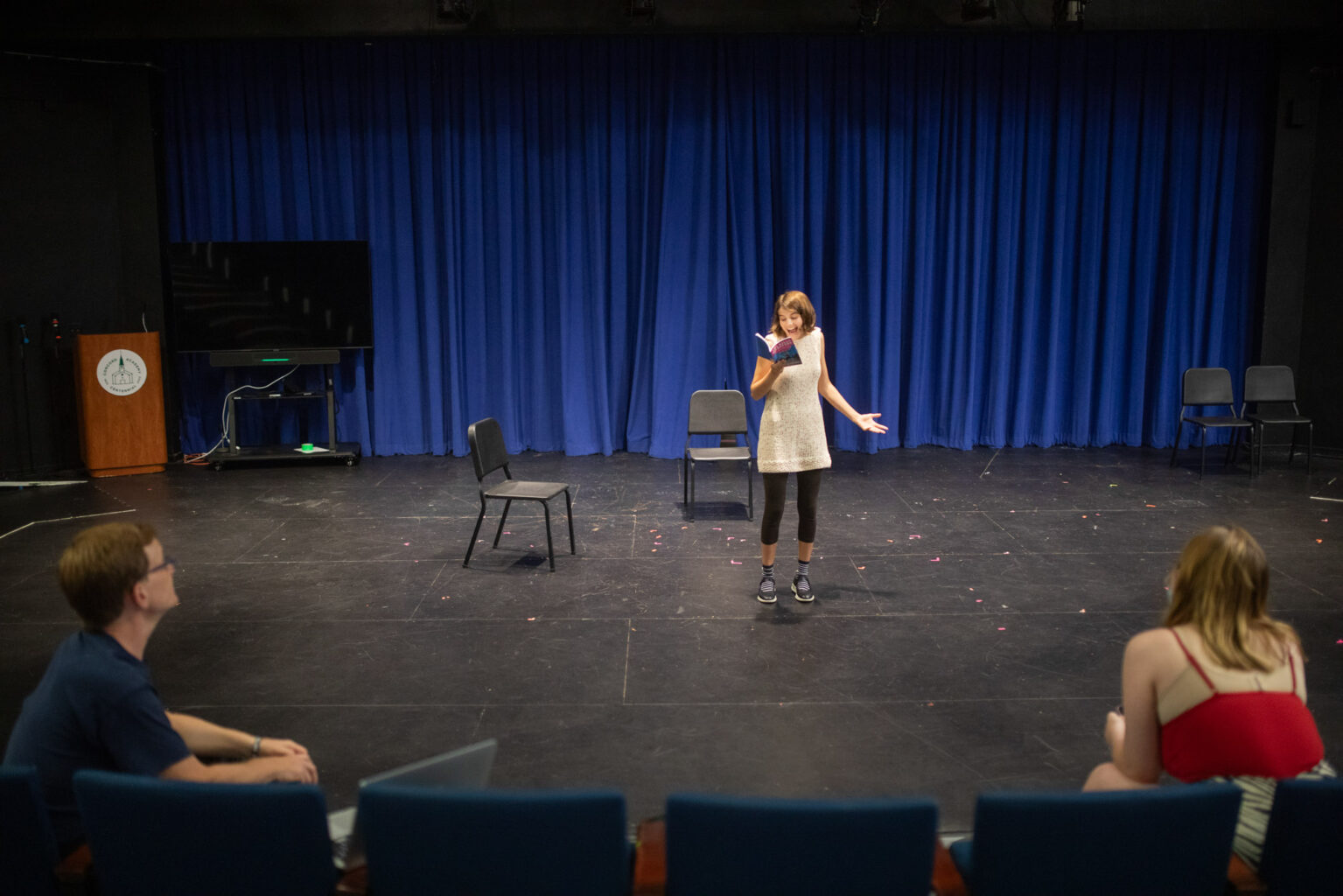 A person stands on a stage speaking and gesturing, with three chairs behind them and two people seated off-stage watching. A blue curtain hangs in the background.