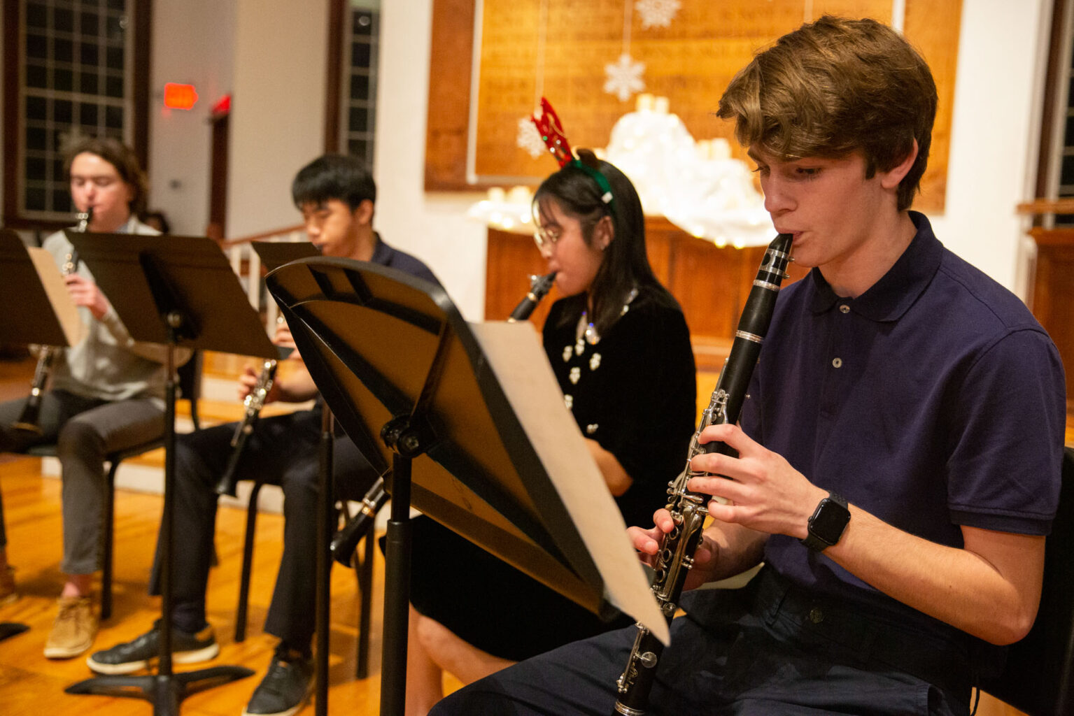 A group of four people, seated and playing clarinets, perform music in a room. One person wears reindeer antlers. Music stands are in front of them.
