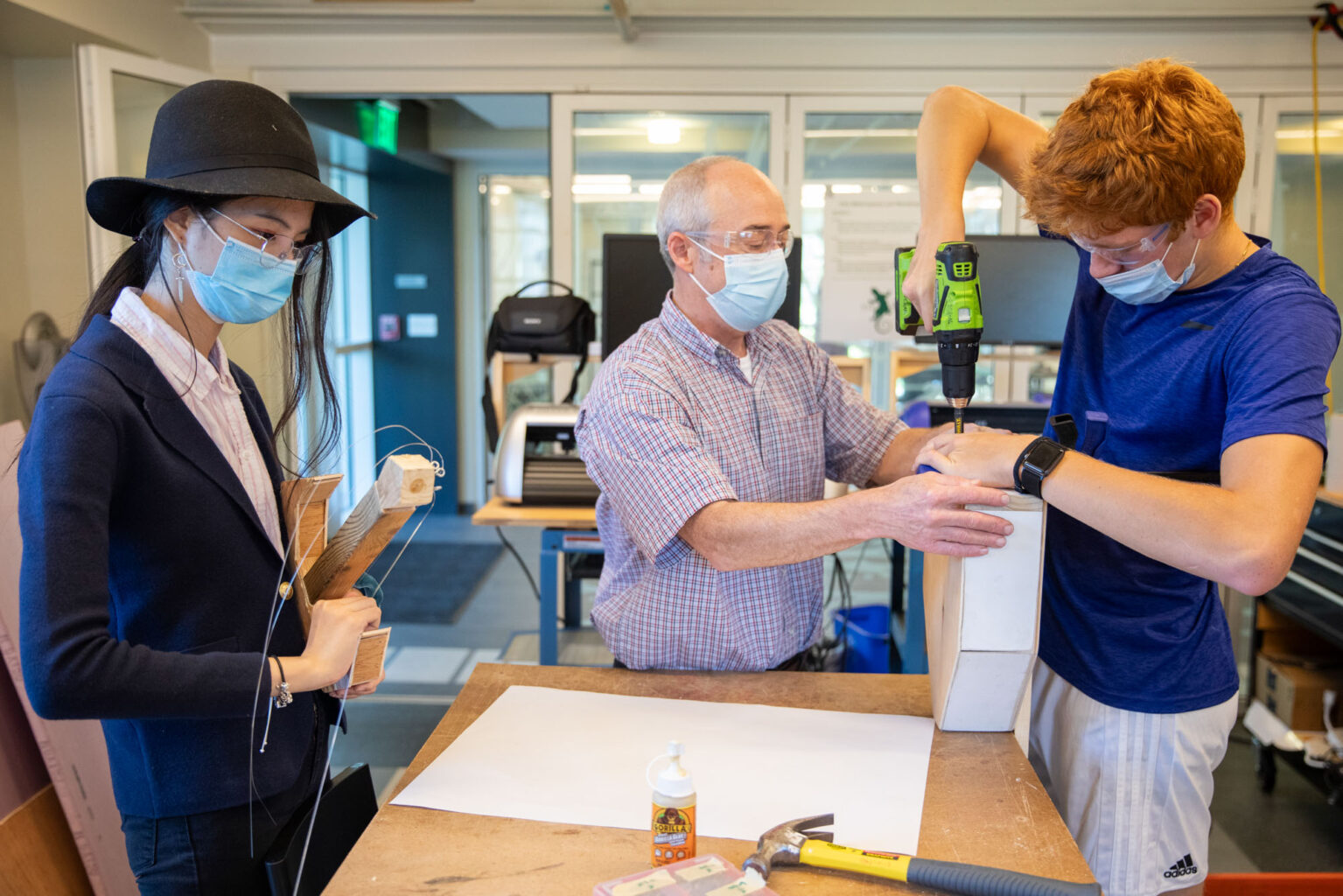 Three people work on a project: one holds wood pieces, another guides a drill, and a third operates it. They wear masks and are in a workshop setting with tools and materials on a table.