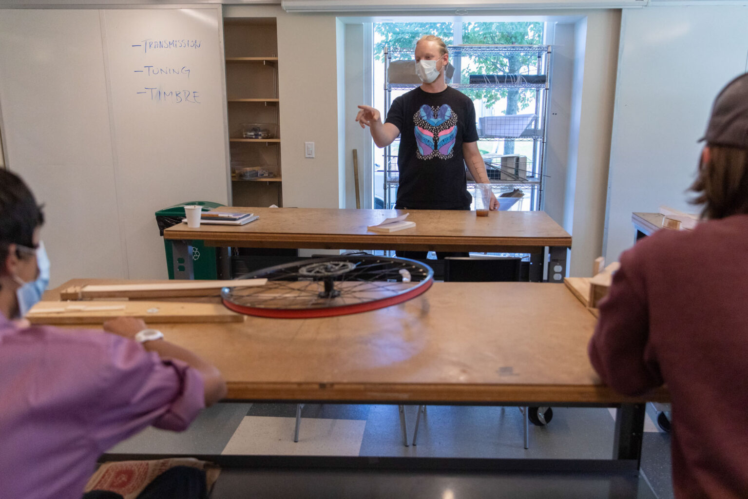 An instructor wearing a mask is standing and gesturing at a table in a classroom while students, also wearing masks, sit and listen. A bicycle wheel lies on a table in the foreground.