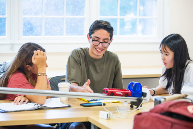 Three students sit at a table in a classroom, collaborating on a project. One student points at a paper while the other two listen attentively. Books and a toy car are on the table.