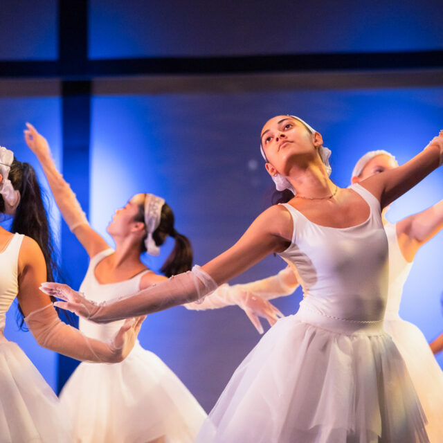 A group of ballet dancers in white costumes perform with graceful arm movements on stage under blue lighting.