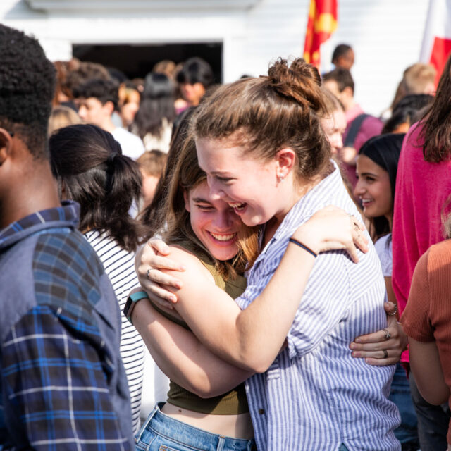Two people embrace and smile while standing amidst a crowd at an outdoor event.