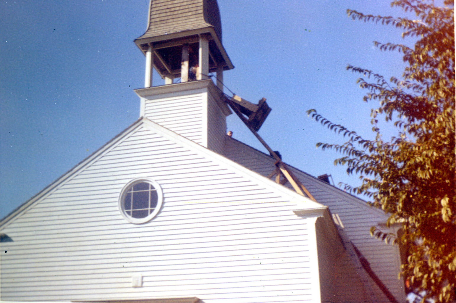 A white church with a damaged steeple under repair; several wooden beams support the steeple. Clear sky and a tree are visible nearby.