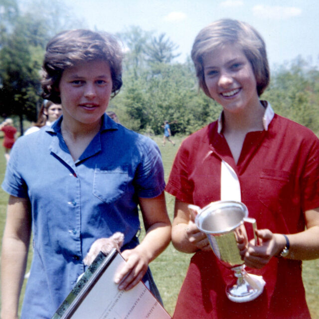 Two young women stand outdoors on grass. The woman on the left holds a book and papers, while the woman on the right, wearing a red shirt, smiles and holds a trophy. Trees and bushes are in the background.