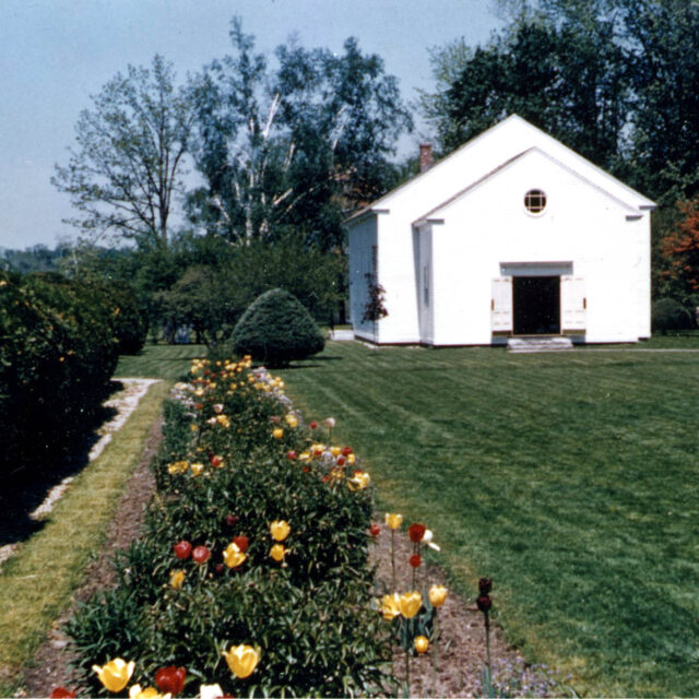 A white house with an open door is surrounded by a green lawn and neat flower beds with blooming tulips, bordered by a hedge with trees in the background.