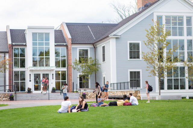A group of students sits on the grassy lawn, with others walking nearby in front of a modern, multi-story building with large windows.
