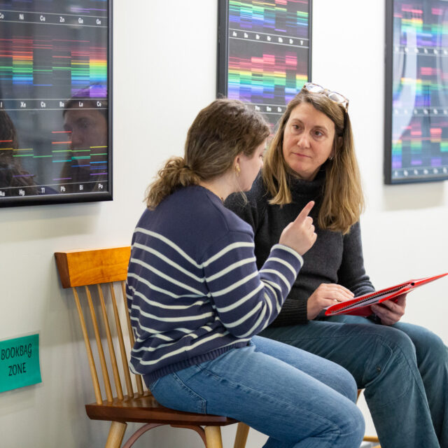Two individuals are seated on wooden chairs in a hallway, engaged in conversation. One person is holding a tablet, and colorful charts hang on the wall behind them.