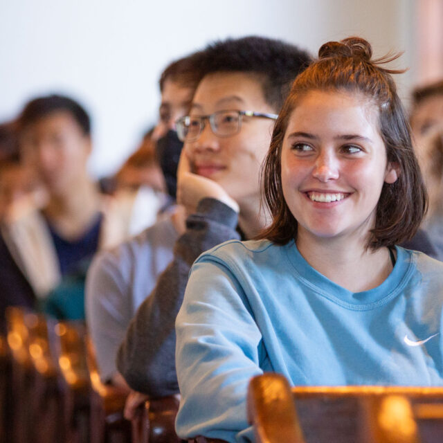 A group of people in casual attire are seated in rows, listening attentively. Some are smiling, and one person in a blue sweatshirt is prominently in focus.