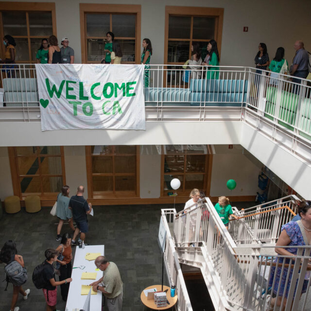 A school lobby with people on both floors. A sign saying "WELCOME TO CA" is hung from the upper railing. People are interacting, and green decorations are seen throughout the area.