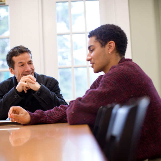 Two people are seated at a wooden table in a room with large windows. They are engaged in conversation, with one person looking at the other attentively while the other speaks.