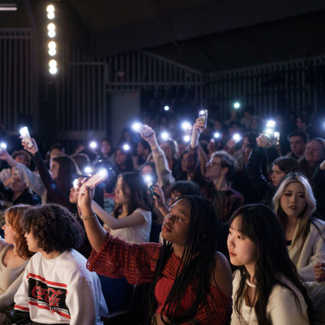A group of people seated in a dimly lit auditorium hold up smartphones with flashlights on, creating a sea of lights.
