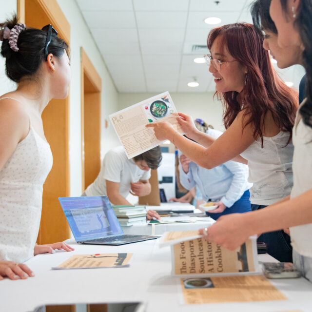 A group of people at a table are discussing documents and using a laptop. One of the women is pointing at a paper while others look on.