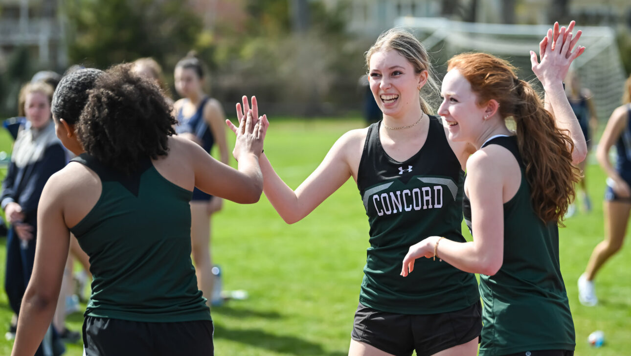 Three young women in green athletic uniforms high-five each other on a track field, celebrating during a sports event. Other athletes and spectators are visible in the background.