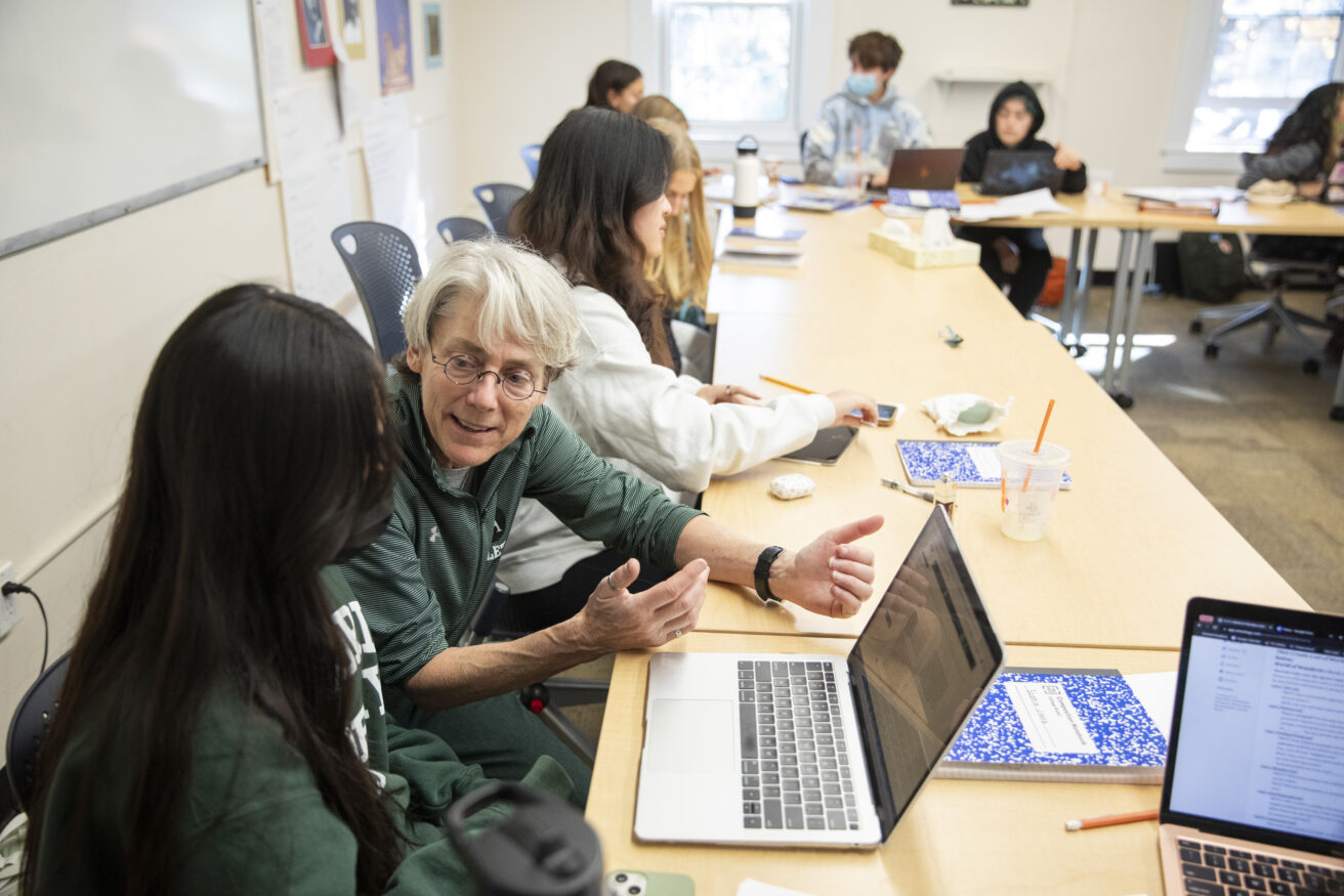 A gray-haired female teacher sits with a student at a table, gesturing to an open laptop.