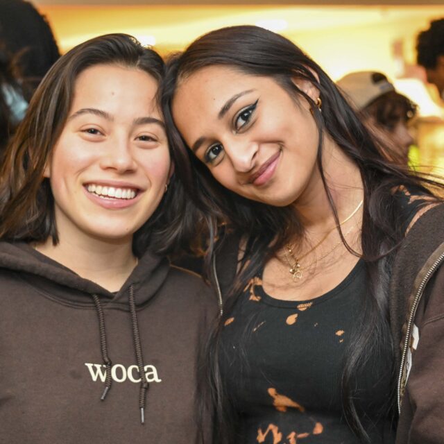 Two girls smile for the camera at a social event; one wears a sweatshirt that says WoCA, which stands for Women of Color Alliance.